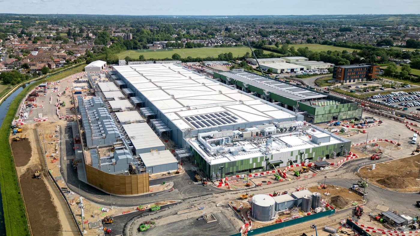 An aerial view of a large data centre campus in daylight, long halls with rooftop plant and cooling equipment, its perimeter road and compound below