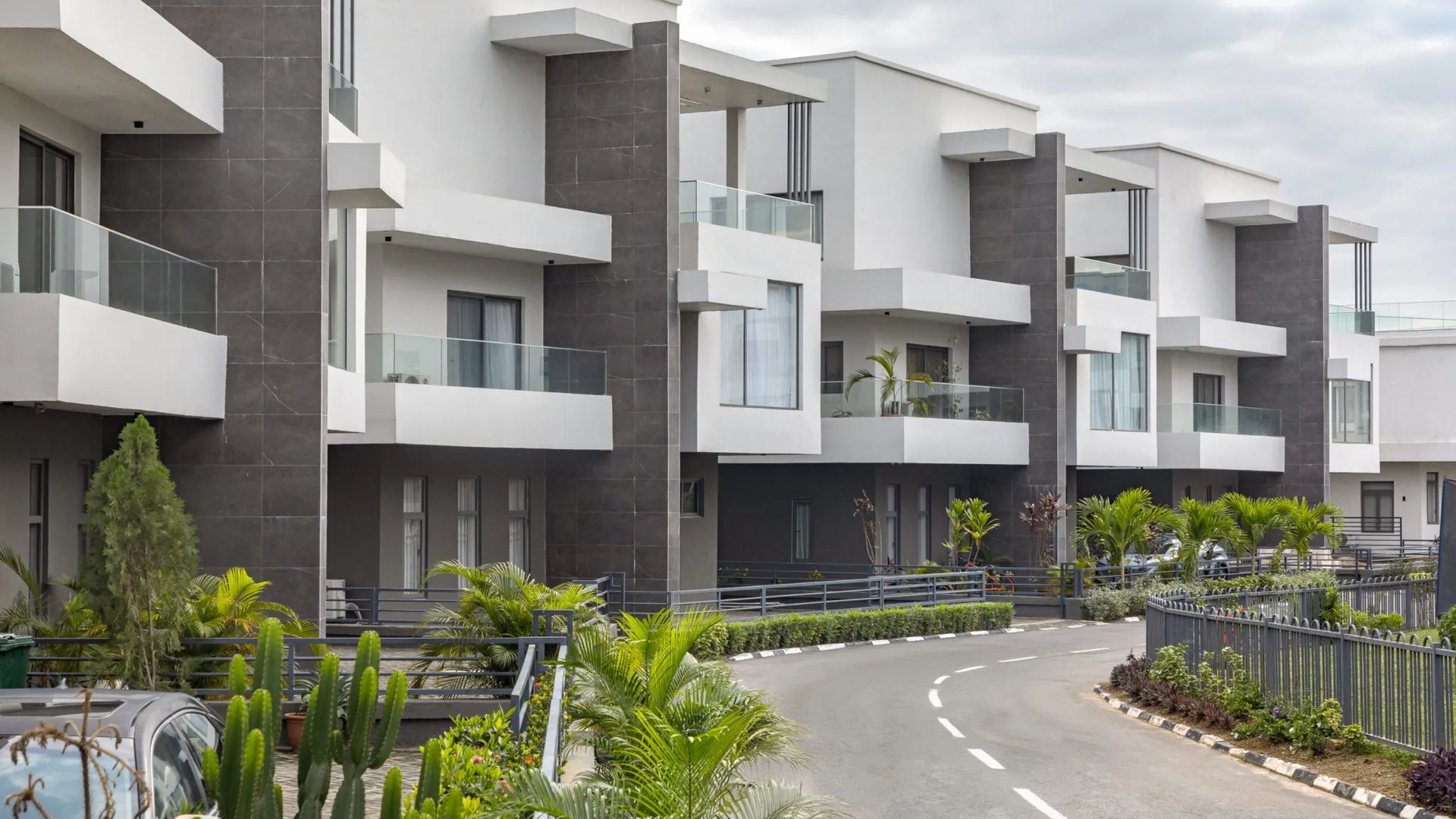 A terrace of modern estate houses along an internal road in daylight, with railings, planting and a perimeter fence