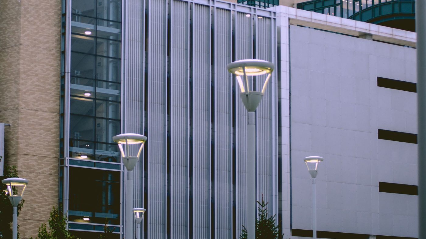 A hospital campus at dusk: a glazed stair tower, lamp posts along the approach and a multi-storey car park behind