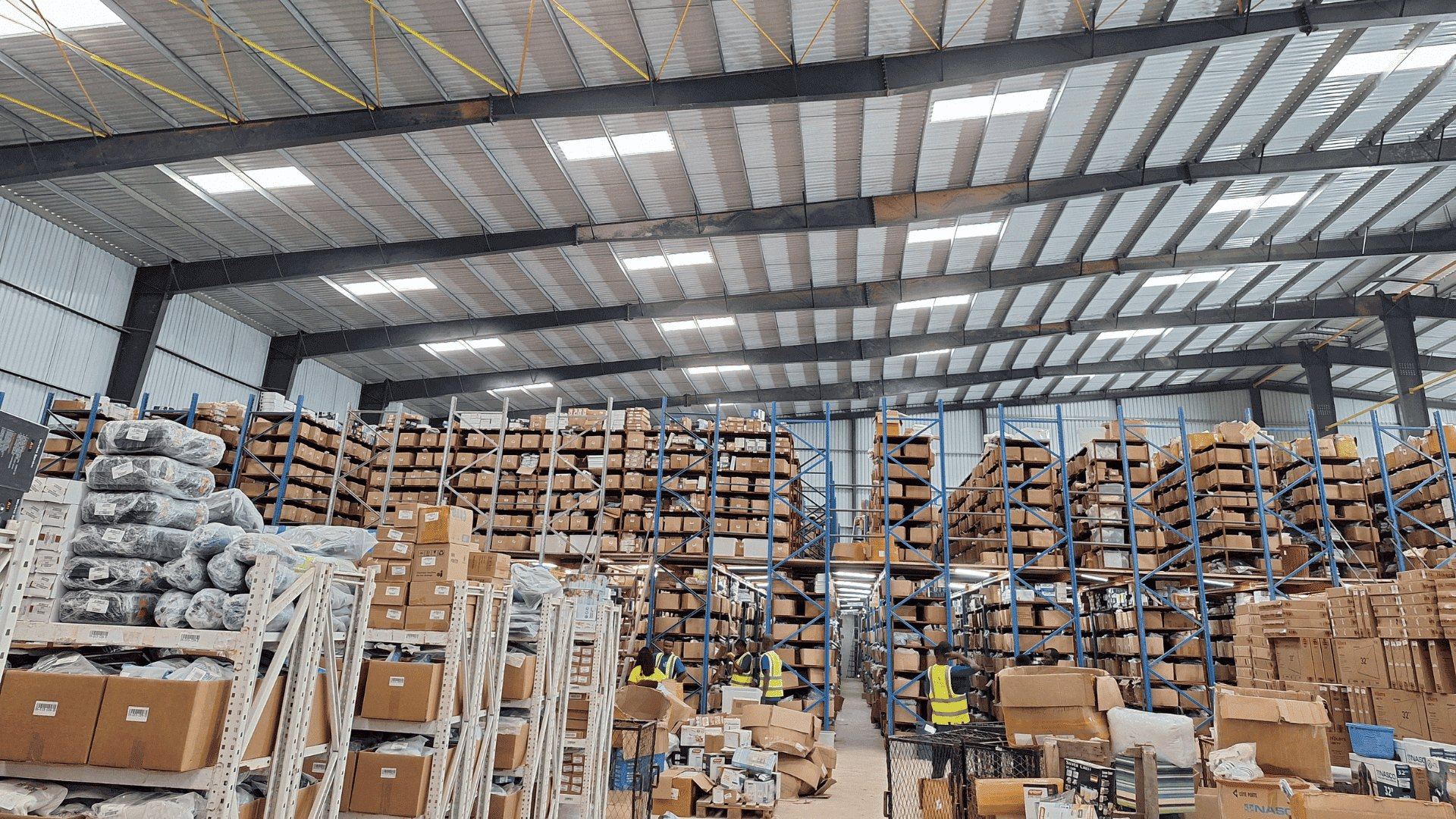 Inside a distribution warehouse: pallet racking stacked to the roof, packed cartons on the floor, and staff in hi-vis working the aisles