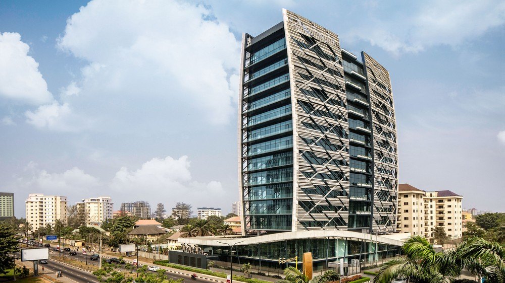 A curved glass and concrete office tower in Lagos seen from across the road in daylight, its glazed ground-floor lobby open to the street