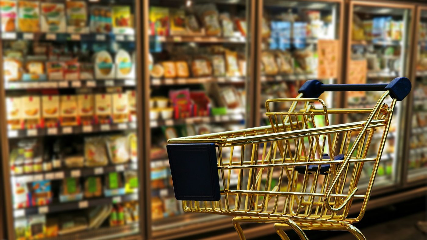 A supermarket aisle in daylight, chilled cabinets stacked with product behind an empty trolley in the foreground