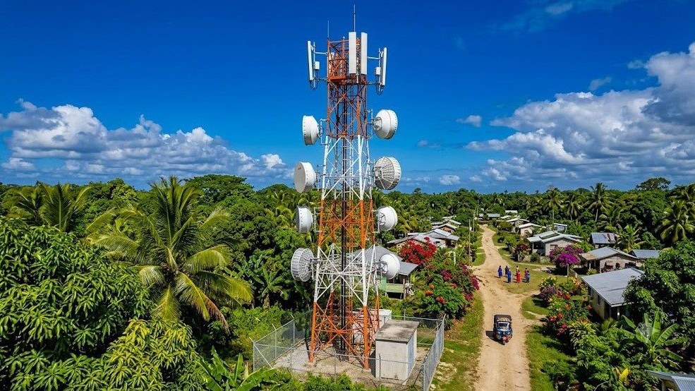 A lattice telecom mast carrying antennas and microwave dishes above a rural village, its equipment cabinets inside a fenced compound at the base